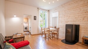 Een gezellige woonkamer met een houten vloer, een bank, een eettafel met stoelen, een zwarte tv-kast, en een keuken met een wasmachine.