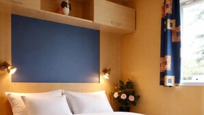 Un dormitorio con cama, mesita de noche, estante en la pared, ventana con cortinas y lámpara de pared.