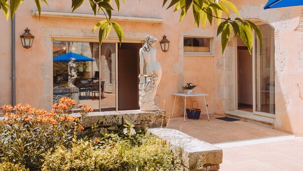 A building with a statue in front, a table with a basket, and a blue umbrella. - Lovely french house in a medieval village - private pool, ping pong & game room (Vianne)