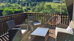 Une terrasse en bois, agrémentée de meubles d’extérieur, notamment des fauteuils en osier et une table, qui offre une vue imprenable sur un paysage verdoyant avec des collines et un ciel dégagé.