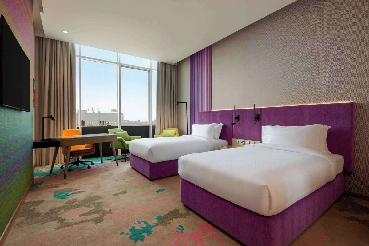 A hotel room with two beds, a desk, a chair, and a large window.