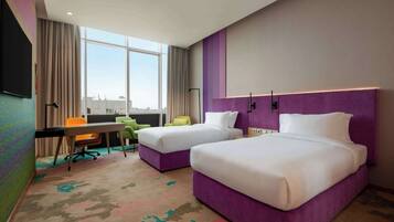 Quarto de hotel com duas camas, uma escrivaninha, uma cadeira e uma janela grande.