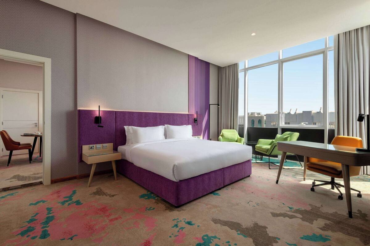 A hotel room with a large bed, a desk, and a chair. There is a window with a view of buildings.