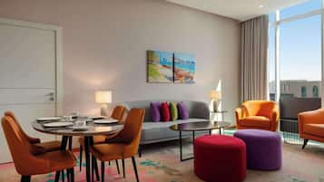 Une chambre d’hôtel moderne comprenant un coin repas, un canapé, deux poufs et un tableau au mur.