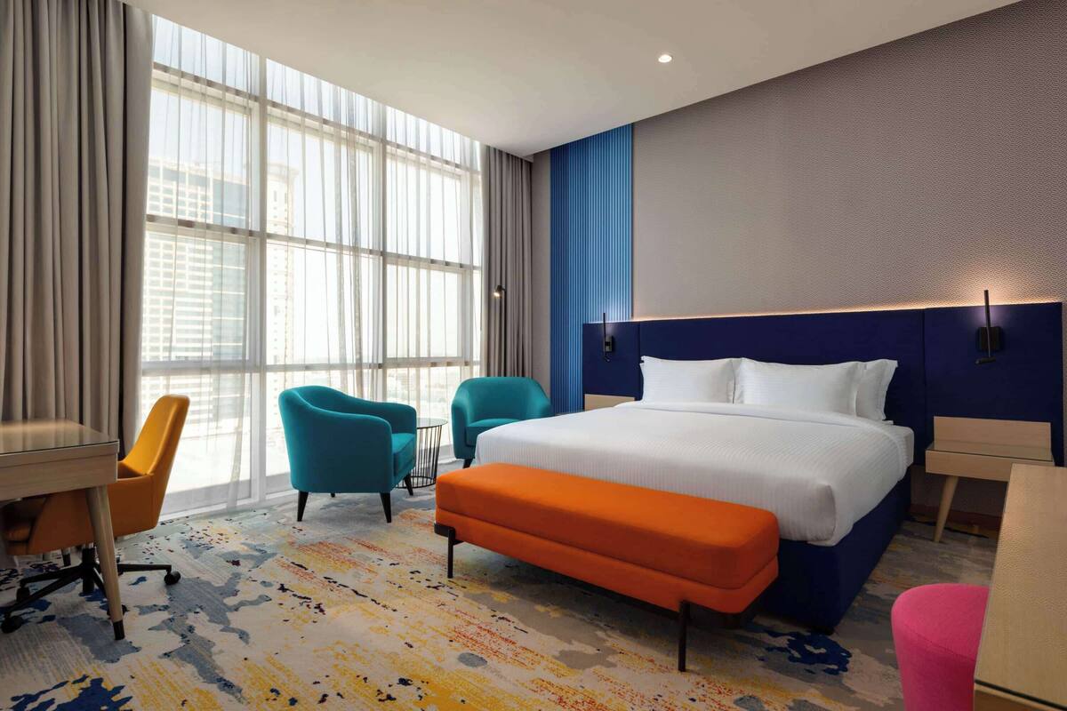 A hotel room with a large bed, a desk, two chairs, and a view of the city through large windows.