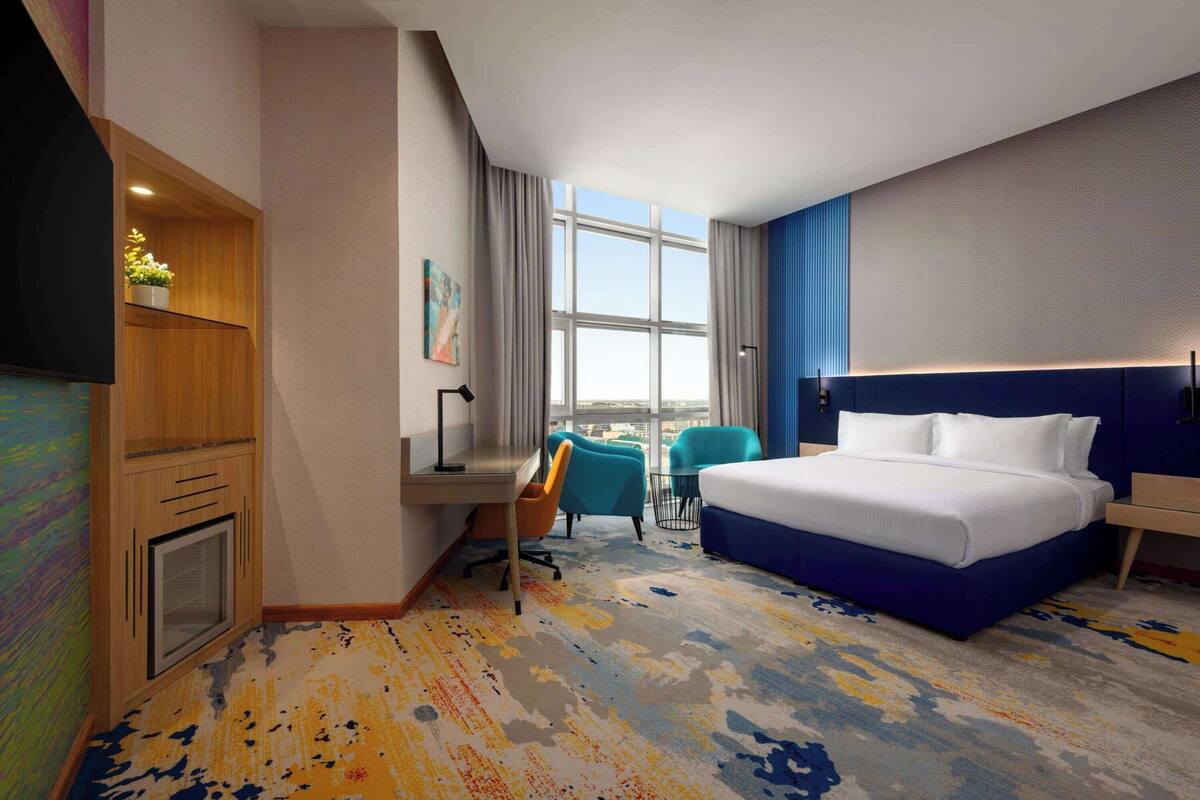 A hotel room with a large bed, a desk, a chair, and a view of the city through a large window.