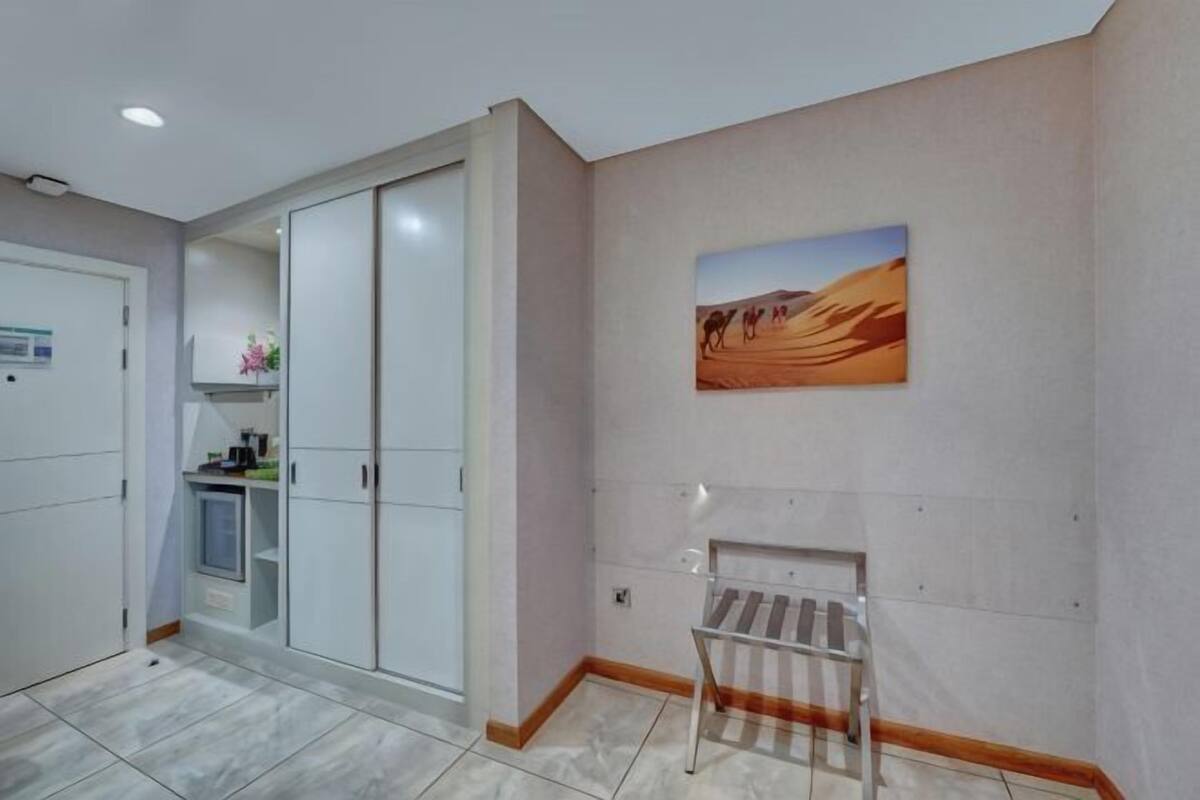 A hotel room with a wardrobe, a painting of a desert, and a bench.