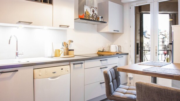 A modern kitchen with white cabinets, a built-in dishwasher, a sink, and a dining area with a table and two chairs.