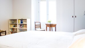 A bedroom with a large bed, a bookshelf, a chair, a small table with a vase, and a window with curtains.
