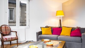 A living room with a grey sofa, colorful cushions, a wooden chair, a glass coffee table with bowls, and a floor lamp.