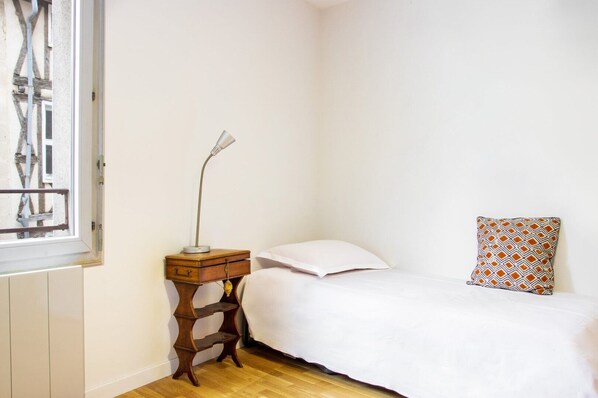 A small, clean room with a single bed, a bedside table with a lamp, and a patterned pillow.
