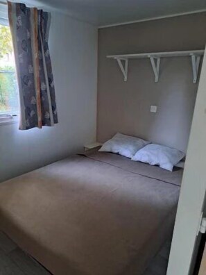 Un lit simple avec une tête de lit, une fenêtre avec des rideaux et une étagère fixée au mur.