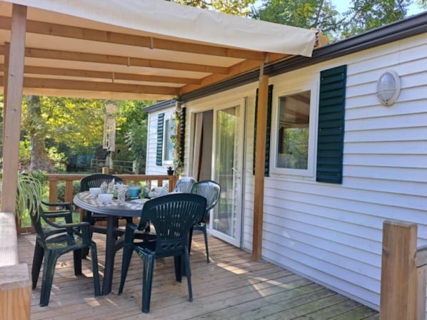 A covered patio with a table and chairs, a wooden deck, and a white building with green shutters.