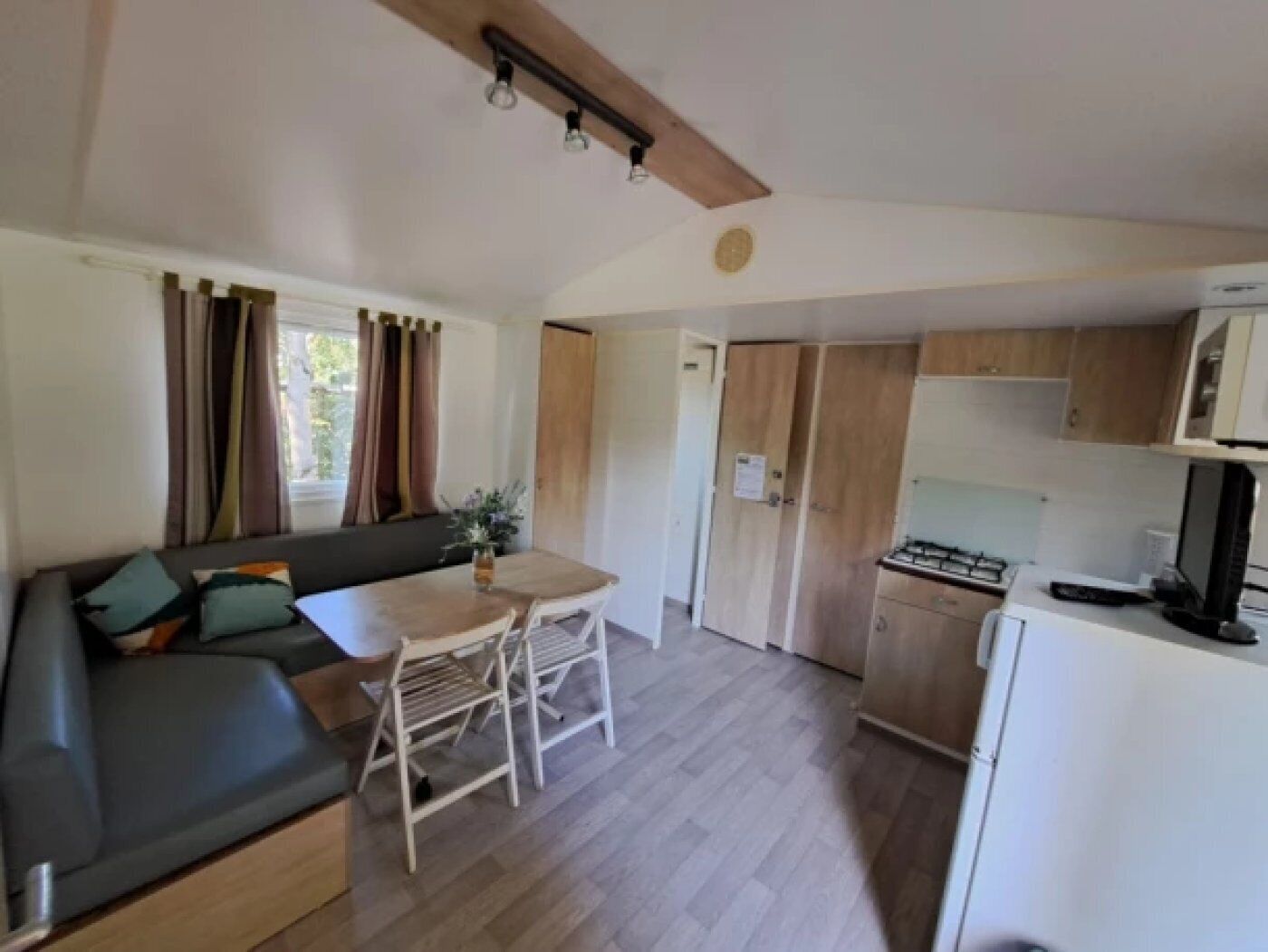 Ein kompakter Wohnbereich mit Sofa, Esstisch, Stühlen, einer Küchenzeile und einem Fenster mit Vorhängen.