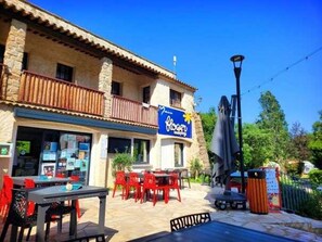 Un restaurant doté d’une terrasse, d’une façade en pierre et d’une enseigne bleue ornée d’un logo représentant un soleil et une fleur.