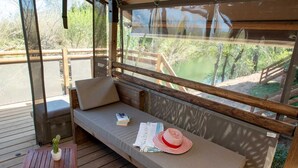 A wooden deck with a cushioned seating area, a small table with a potted plant, and a view of a natural landscape through a window. - ZEcamping 3 étoiles piscine chauffée locatif TV clim 34m2 (Roquebrune-sur-Argens)