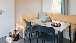 Une petite pièce agréable et chaleureuse, avec un canapé, une table et des chaises. Il y a une fenêtre avec des rideaux, une porte et un panier rempli d’objets posé sur le sol.