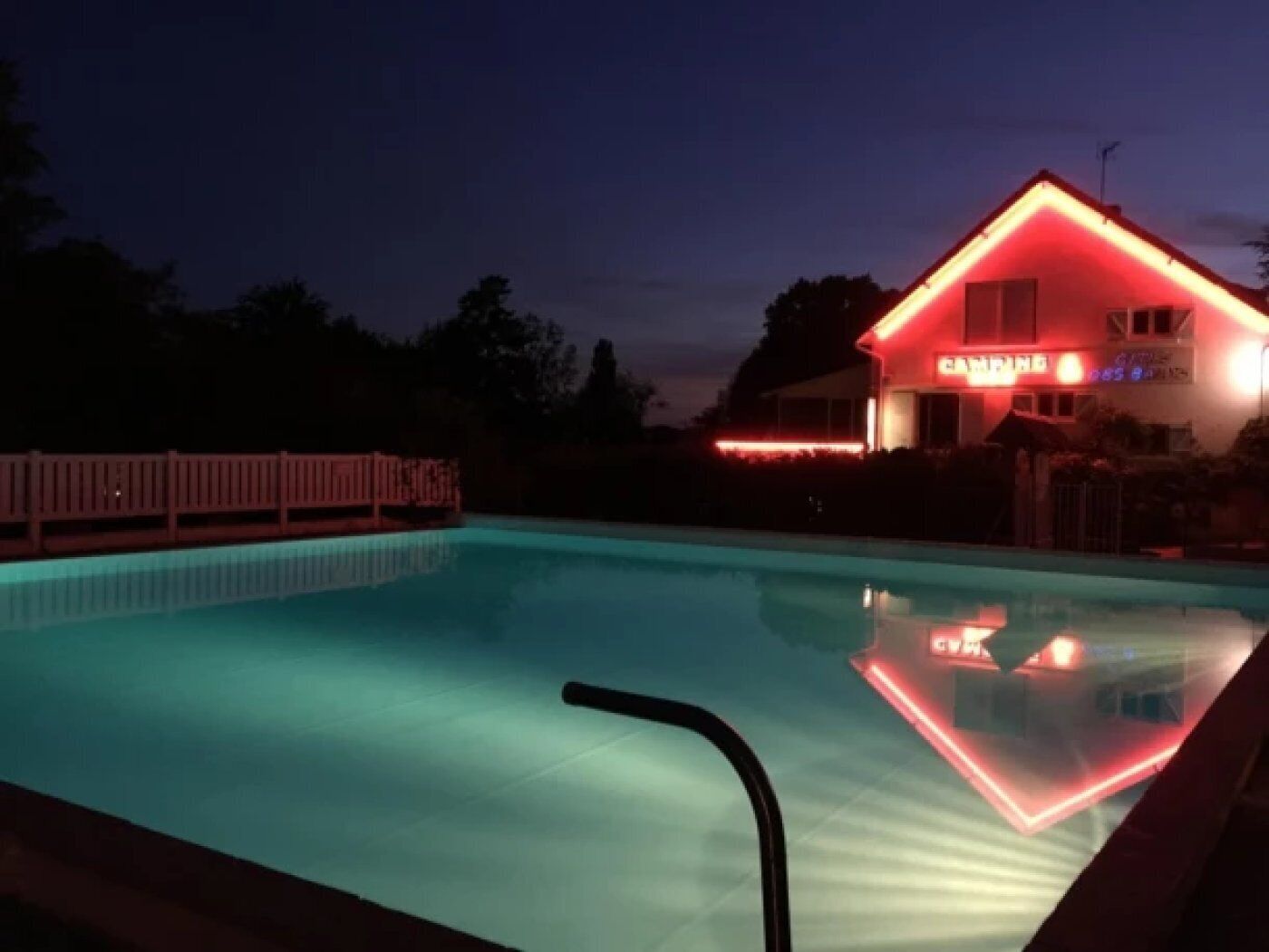 Een zwembad met een houten terras en een huis met rode verlichting op de achtergrond.