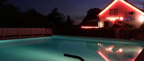 Una piscina con cubierta de madera y una casa con iluminación roja al fondo.
