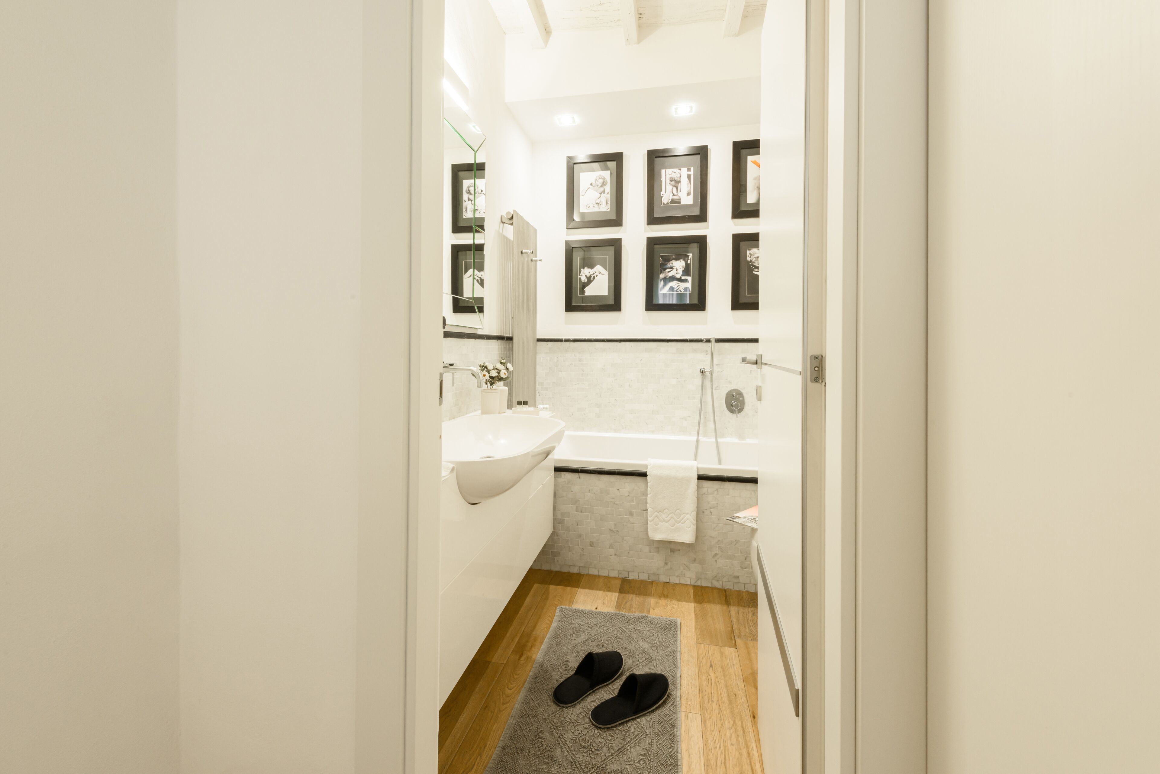 Une salle de bain moderne avec un lavabo blanc, une baignoire et une douche. Des tableaux encadrés ornent le mur et une paire de chaussons est posée sur le sol.