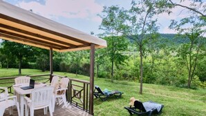Una terraza techada al aire libre con sillas y una mesa blancos, con vistas a una zona verde con árboles y una colina distante.