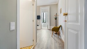 A hallway with a light wooden floor, white walls, and a green chair. There is a door with a gold handle on the right and a light switch on the left. - GuestReady - Alegria 293 - Room 4 (Porto)