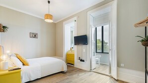 A bedroom with a bed, a yellow nightstand, a woven rug, a television on the wall, and a window with a view. - GuestReady - Alegria 293 - Room 4 (Porto)