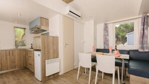 Eine kleine Küche mit Holzschränken, einem weißen Kühlschrank und einem eingebauten Herd. Es gibt einen Essbereich mit Tisch und Stühlen sowie eine Sitzecke mit Blick ins Freie durch ein Fenster.