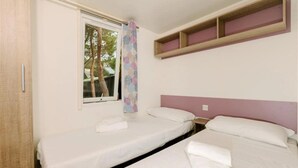 A room with two single beds, a window with a curtain, a wooden shelf above the beds, and a door on the left. - ZEcamping 4 étoiles animé pataugeoire locatif TV clim 41m2 4 pers (Alghero)