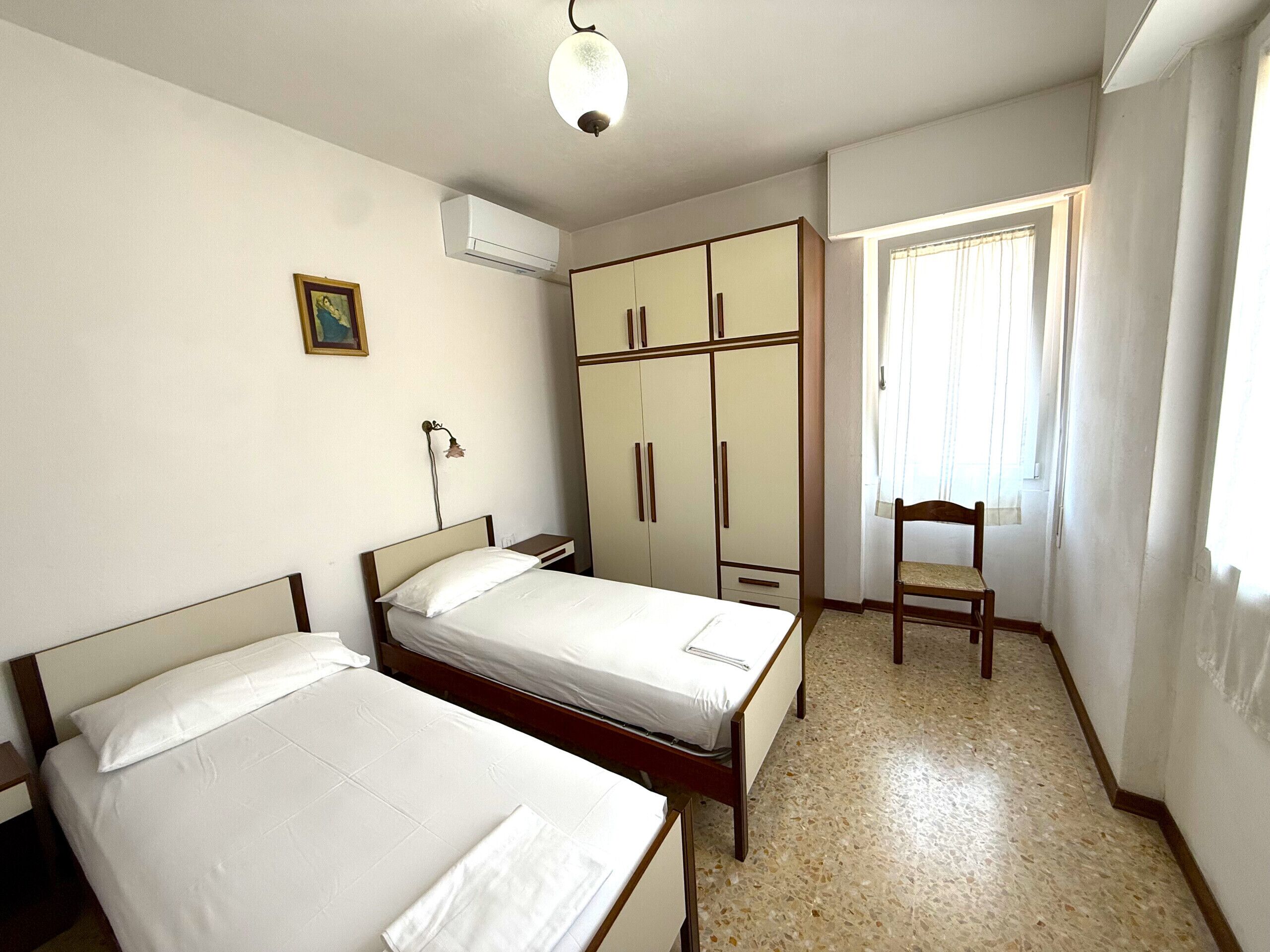A bedroom with two beds, a wardrobe, a chair, and a window with curtains.