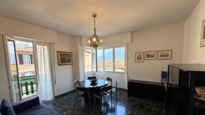 A room with a dining table, chairs, a sofa, a TV, and a view of buildings through the window. - Casa Liver Pieve balcony town center (Tremosine)