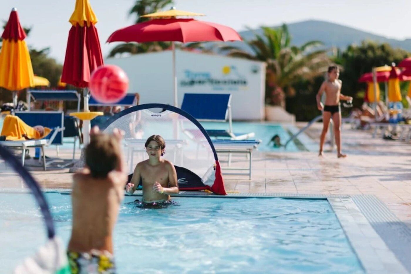 A swimming pool with lounge chairs and umbrellas, a person in the water, and another person standing by the pool.