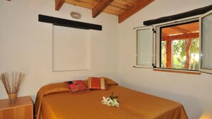 A bedroom with a wooden ceiling, a bed with a brown bedspread, a window with a view of trees, and a bedside table with a vase of reeds.