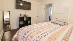 A bedroom with a large bed, a flat-screen TV mounted on the wall, a bedside table with a lamp, and a door leading to another room. - Fun hangout; Perfect Location; Walk to Coliseum (Greensboro)