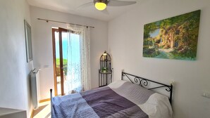 Un dormitorio con cama, ventana con cortinas, ventilador de techo, lámpara de pared y un cuadro.