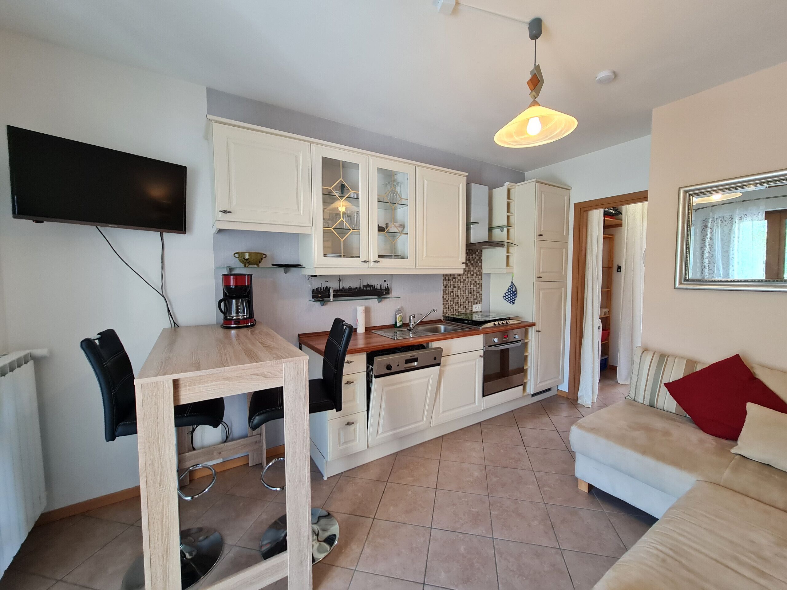 A compact kitchen with white cabinets, a dining table with chairs, a coffee maker, and a TV mounted on the wall.