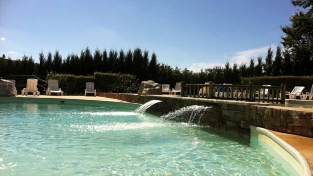 A clear swimming pool with a waterfall feature, surrounded by lounge chairs and a stone wall.