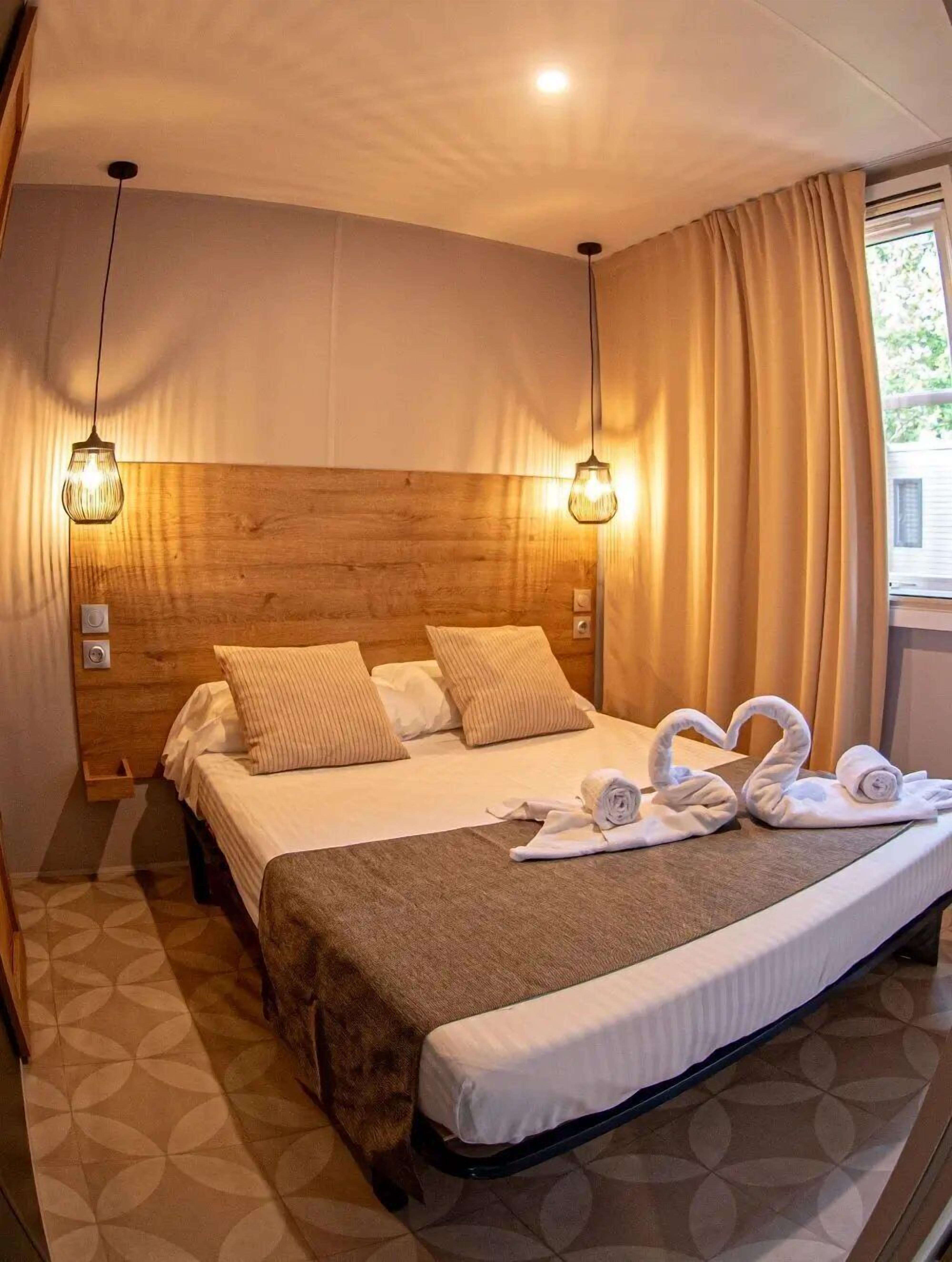 Una habitación de hotel con cabecera de madera, una cama grande con ropa blanca, dos lámparas colgantes fijadas a la pared y una ventana con cortinas.