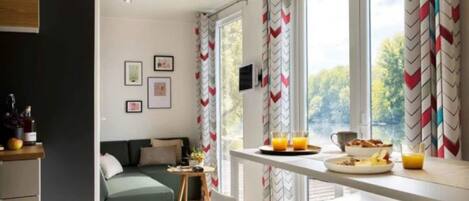 A modern room with a green sofa, a dining table with breakfast items, and large windows with chevron-patterned curtains.