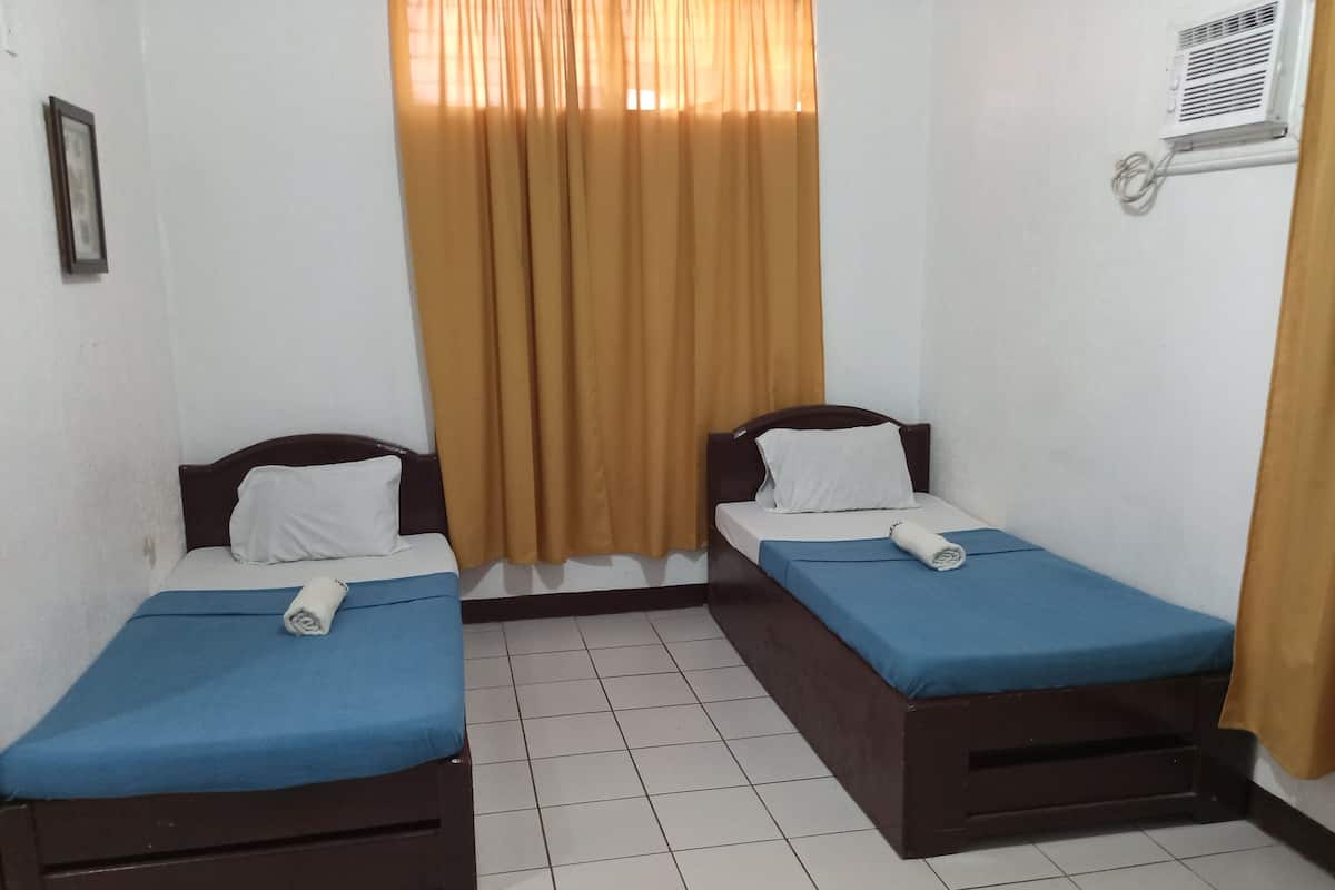 A small, clean room with two single beds, a window with curtains, a wall-mounted air conditioning unit, and a tiled floor.