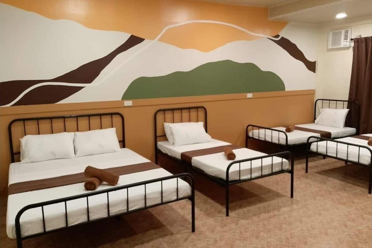 A hotel room with four single beds, each with a metal frame and white bedding. The room features a mural on the wall depicting a landscape.