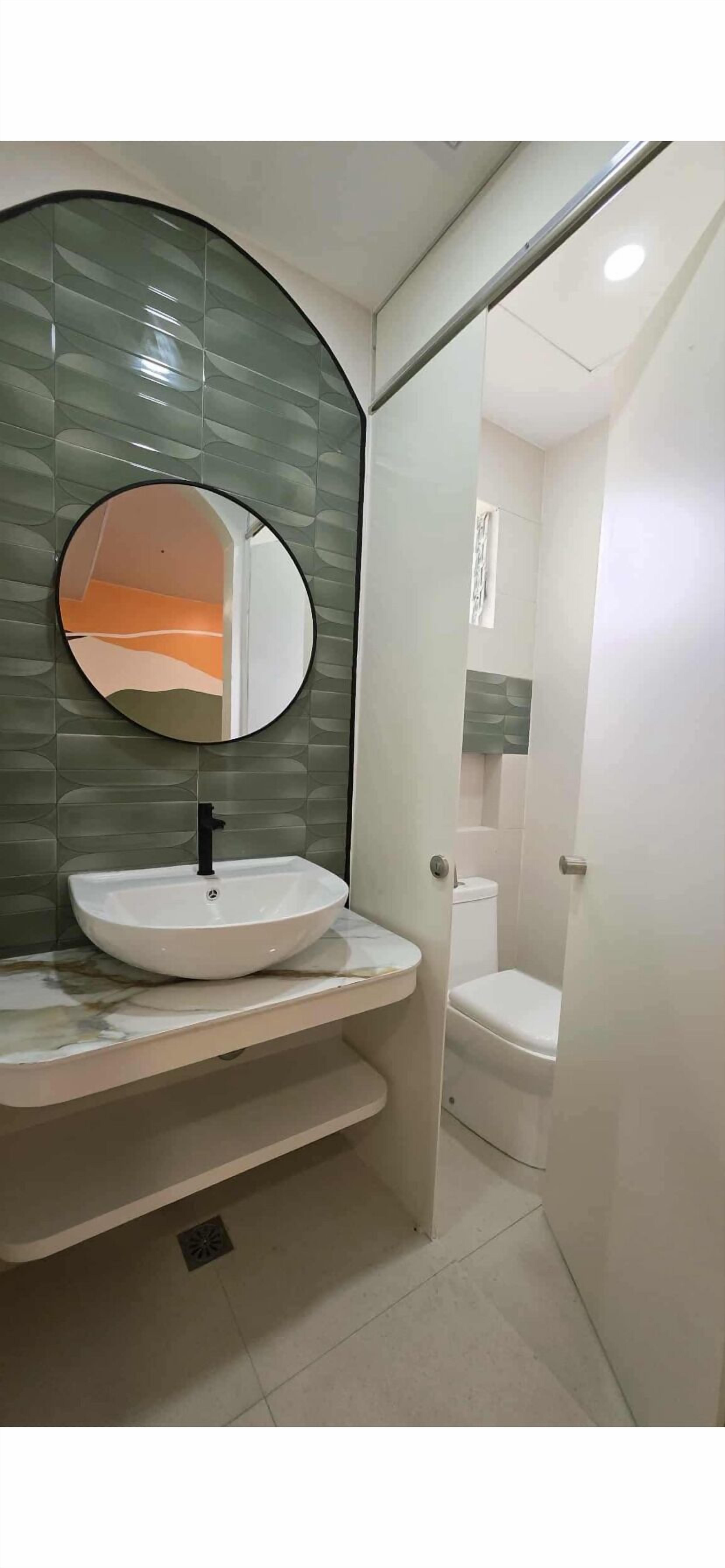 Un baño con un espejo redondo, un lavabo blanco y un inodoro visible a través de una puerta abierta.