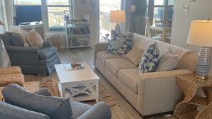 Een woonkamer met een bank, fauteuils, een salontafel en een televisie. Een boekenkast en een eethoek zijn zichtbaar door de glazen deuren.