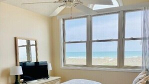 Een slaapkamer met een bed, een plafondventilator, een televisie, een raam met uitzicht op het strand en een dressoir met een lamp.