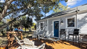 A white house with a blue door, a deck with Adirondack chairs, and a large tree. - Walk to Sound + Dining | Fenced Yard | Extended Stays Welcome Pet Friendly (Morehead City)
