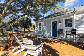 A white house with a blue door, a deck with Adirondack chairs, and a large tree.
