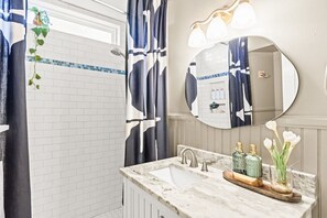 A bathroom with a white tiled wall, a sink with a marble countertop, a mirror with a light fixture, and a shower curtain with a black and white pattern.