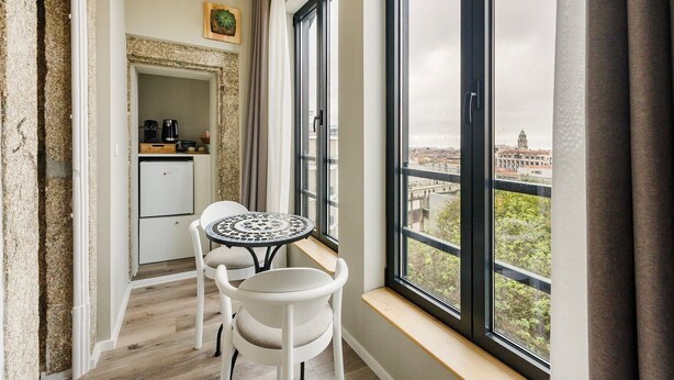 Ein schmaler Balkon mit einem kleinen Tisch und zwei Stühlen, eine Küchenzeile mit einer Kaffeemaschine und Blick auf eine Stadtlandschaft durch große Fenster.