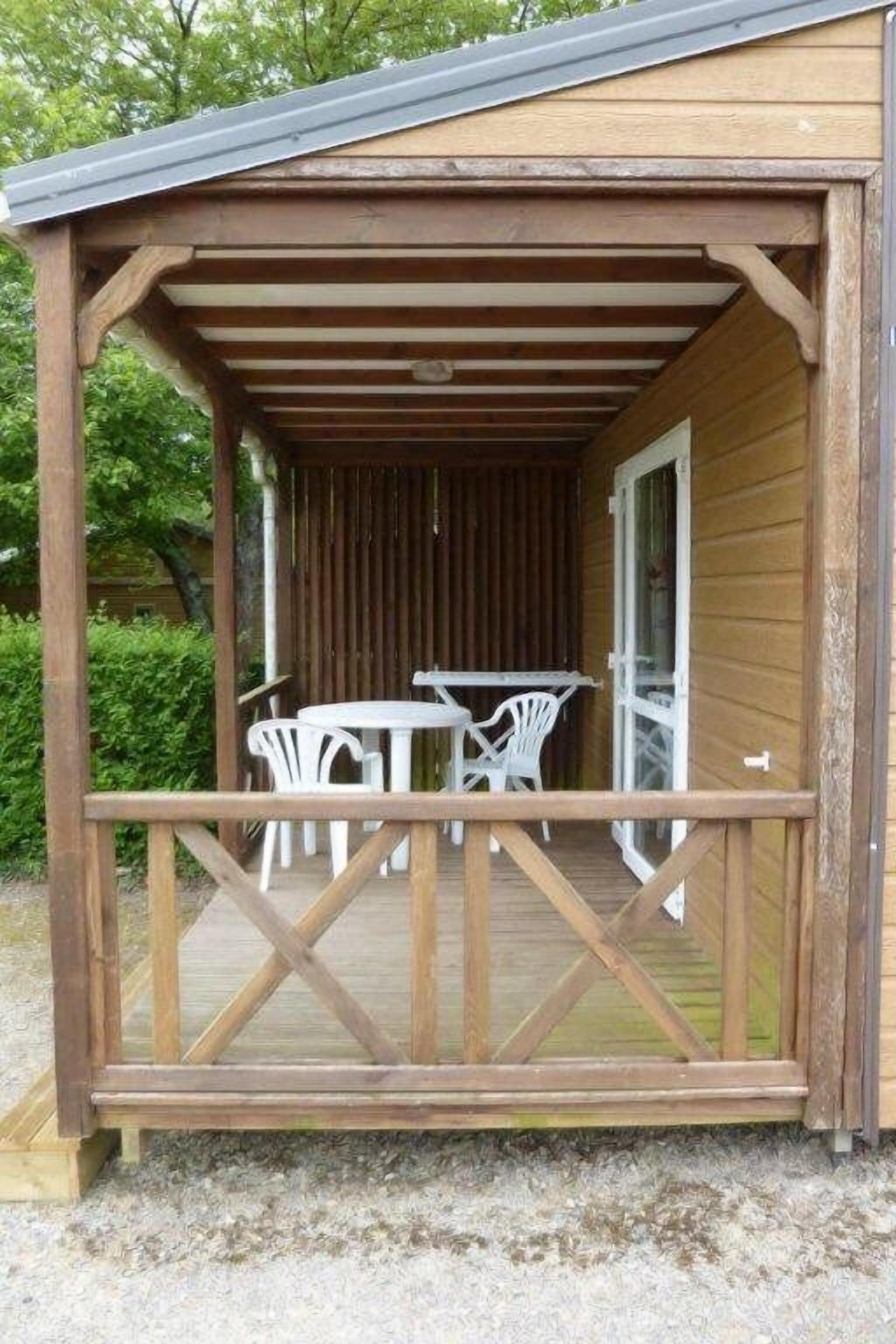 Une terrasse en bois avec une table et des chaises, un espace couvert avec une porte, et une vue sur la verdure en arrière-plan.