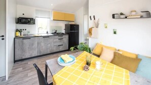 A modern kitchen with grey cabinets, a black refrigerator, and a microwave. There is a dining area with a yellow table and chairs, and a small plant on the windowsill. - ZEcamping 4 étoiles familial 500m plage espace aquatique locatif TV (Benodet)